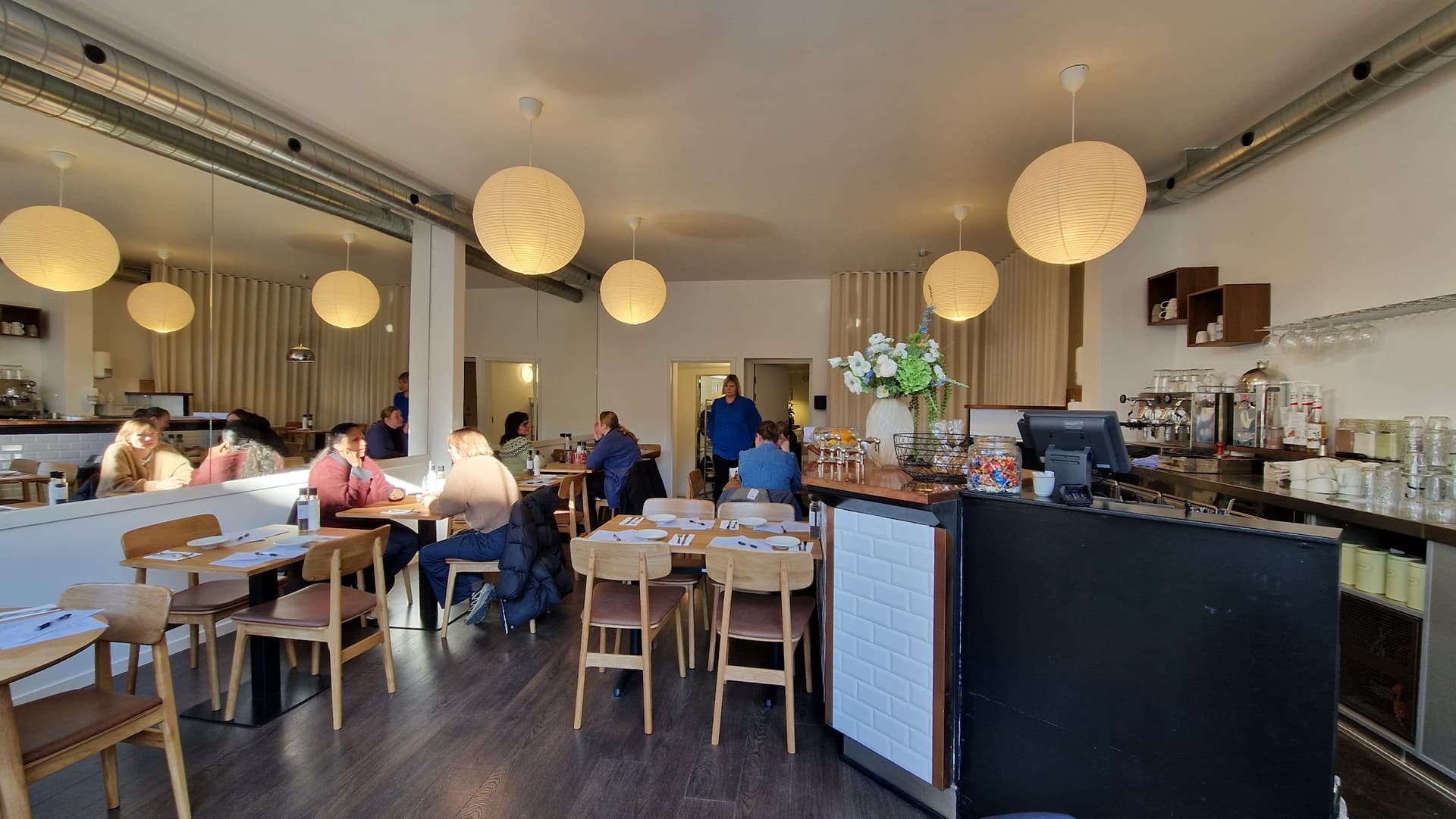 Lyst caféinteriør hos MUNN med hvide papirlamper, egetræsborde og den mørke kaffebar med espressomaskiner